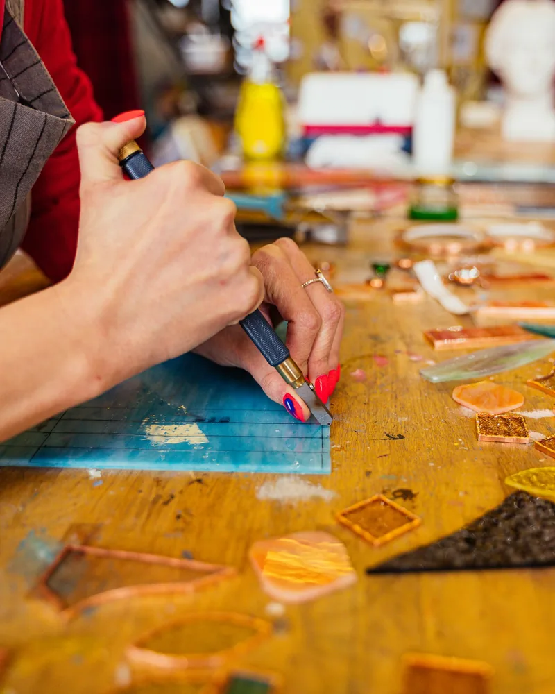 Manos cortando una plancha de vidrio con un cortavidrios sobre una mesa de taller cubierta de recortes de vidrio de colores.